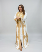 A woman modeling a long white caftan-style dress with extensive gold embroidery and gold trim, standing against a light gray studio background.