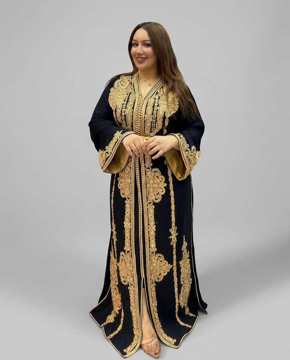 A woman modeling a long black caftan-style dress with extensive gold embroidery and gold trim, standing against a neutral gray studio background.