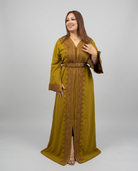 A woman modeling a long olive green kaftan dress with intricate gold embroidery and a matching belt, photographed against a solid light gray studio background.