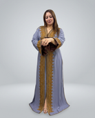 A woman modeling a long, floor-length light blue abaya with ornate gold embroidery and trim, standing against a neutral gray studio background.