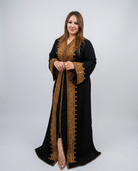 A woman modeling a black long kaftan dress with ornate gold embroidery and a matching inner robe, standing against a light gray studio background.