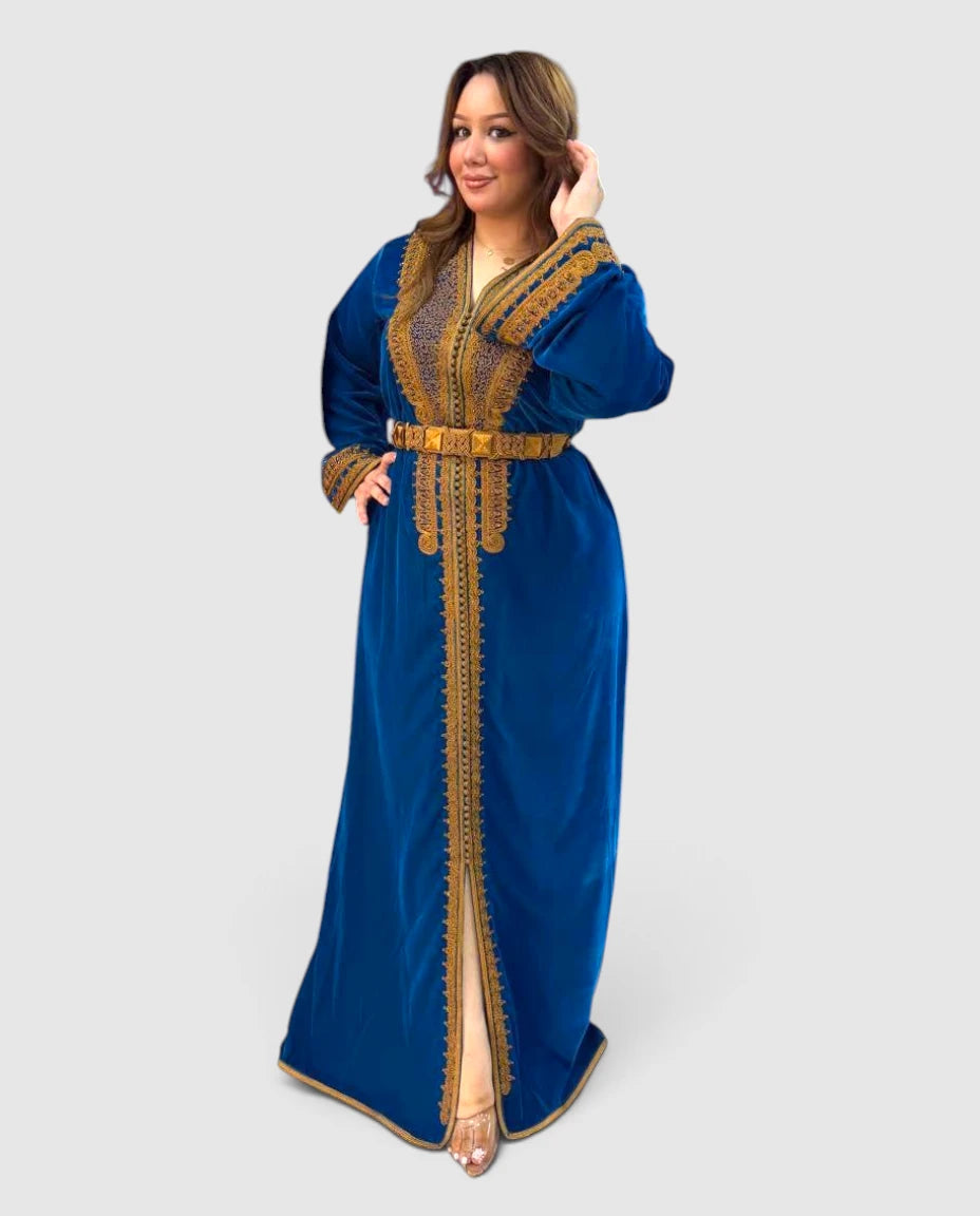A woman modeling a long royal blue velvet caftan dress with ornate gold embroidery, a matching gold belt, and a high front slit, set against a solid light gray background.