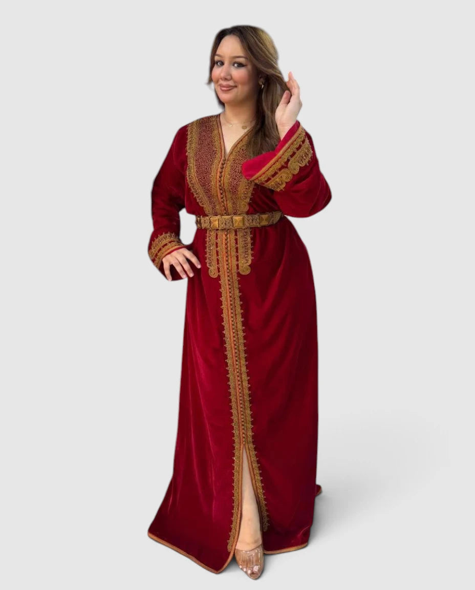 A woman modeling a long red velvet caftan dress with ornate gold embroidery, a matching belt, and a high front slit, set against a solid light gray background.
